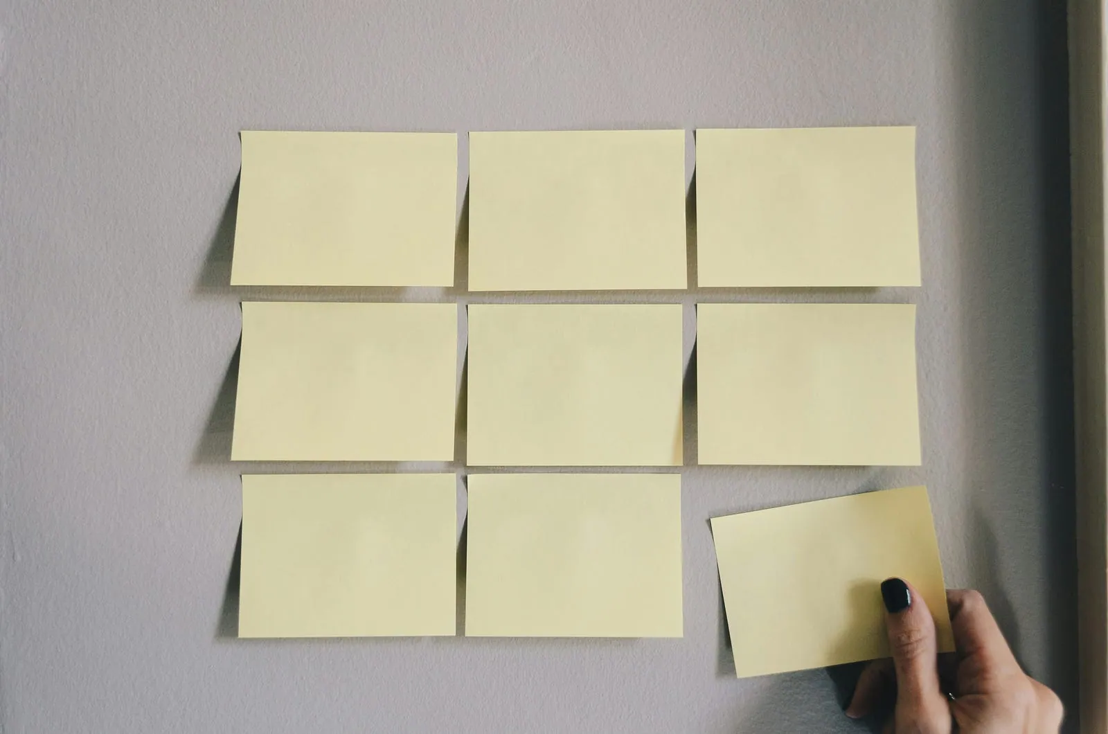 A hand placing the last of nine blank sticky notes in a grid on a wall.